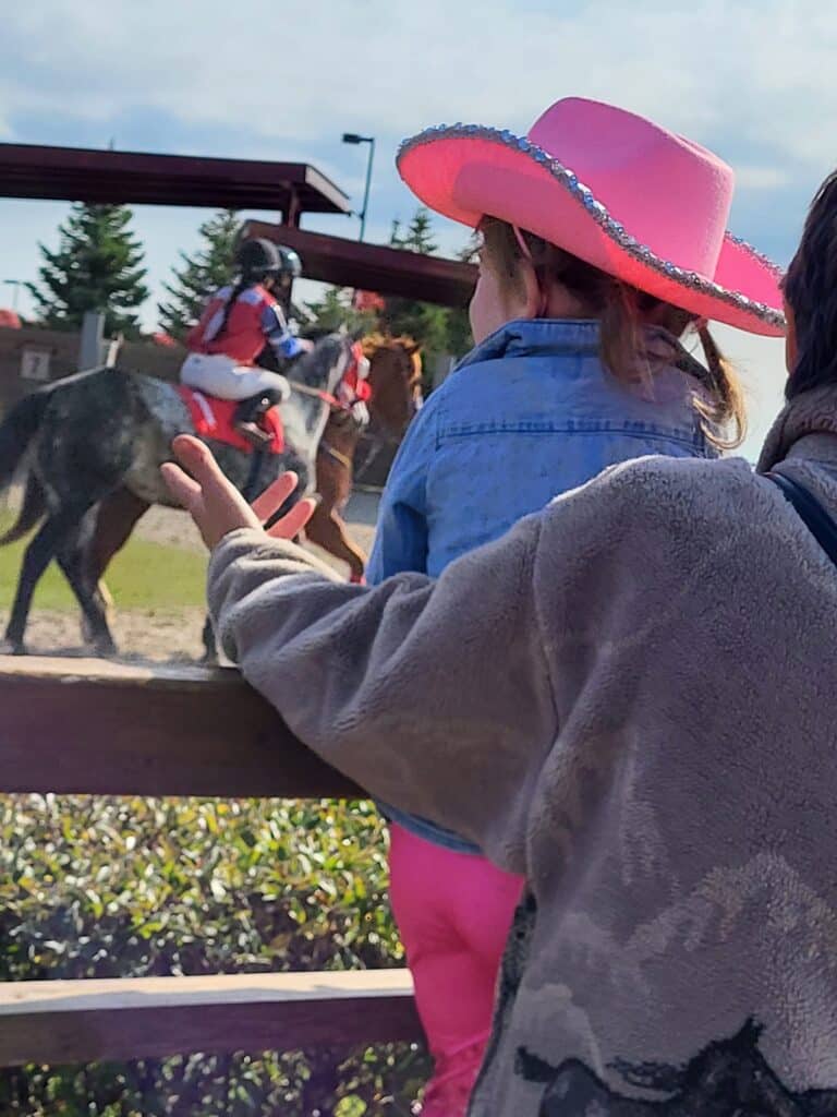 A little girl wearing a pink cowgirl hat, watching some racehorses.