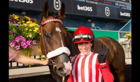 A female jockey in red-and-white silks holding a horse and waving.