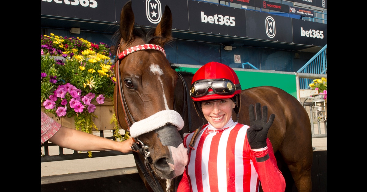 A female jockey in red-and-white silks holding a horse and waving.