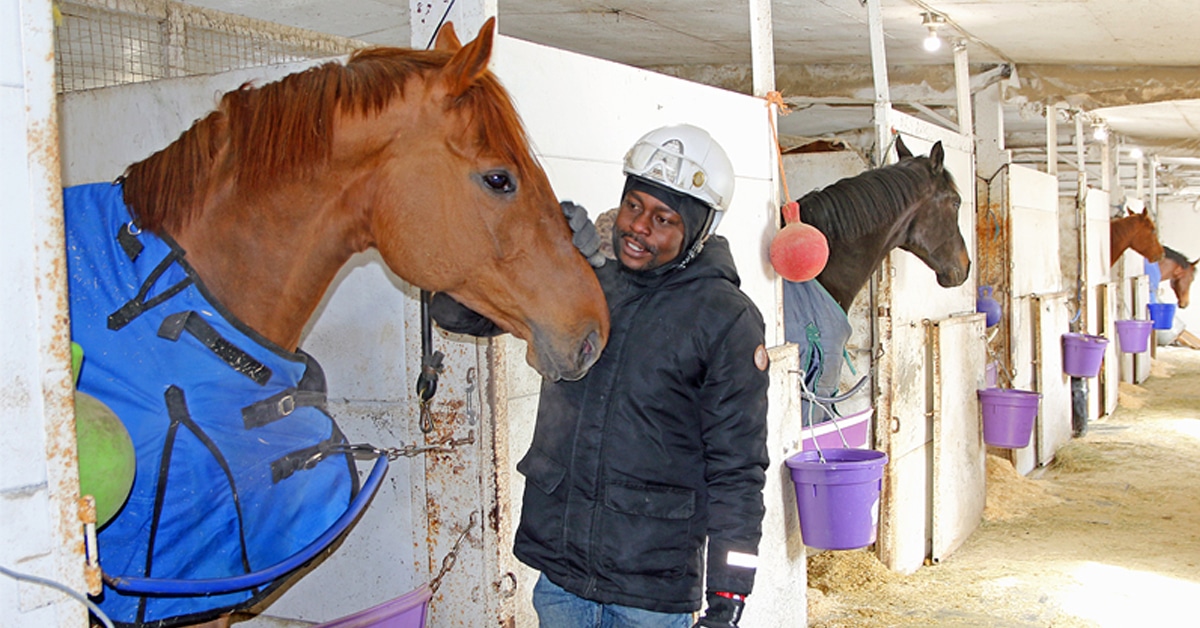A man patting a chestnut horse in a shedrow.